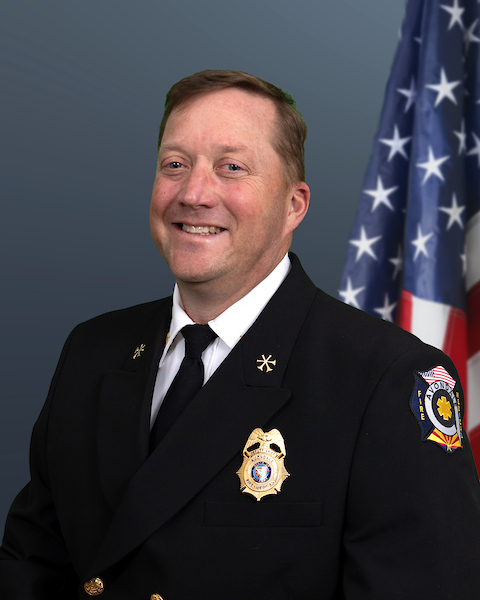 Chief Glass from Avondale Fire & Medical poses for an official portrait in front of an American flag. He is smiling and dressed in a dark formal dress uniform with department maltese on his left shoulder, a gold badge on his left chest, and collar pins indicating rank. The background is a plain gradient blue.
